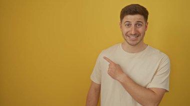 Yakışıklı, İspanyol asıllı, gülümseyen ve izole edilmiş sarı bir arka plan duvarına karşı dururken ekranda olmayan bir şeye işaret eden bir adam..