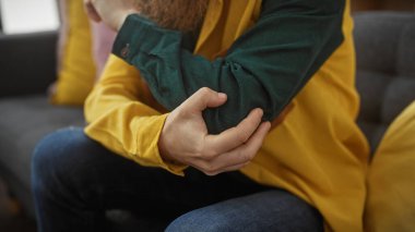 Sarı gömlekli genç bir adam dirseğine dokunuyor, evde acı ya da rahatsızlık belirtileri gösteriyor..