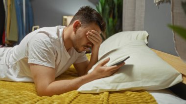 Sakallı genç bir İspanyol adam yatak odasında bir yatakta uzanıyor, akıllı bir telefon tutuyor ve yüzünü eliyle kapatıyor, hüsran ya da stres iletiyor..