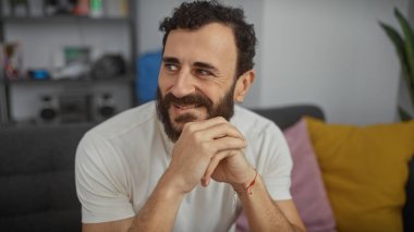 Sakallı, gülümseyen, orta yaşlı bir adam evde gündelik bir ortamda, arka planda odakları bozuk, modern bir oturma odası var..