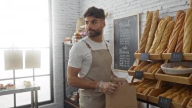 Genç adam bir fırında çalışıyor, bir kese kağıdına taze ekmek dolduruyor, arkasında kırsal bir iç mekan ve çeşitli pastalar sergileniyor..
