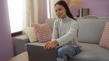 Güzel bir kadın, şık bir oturma odasında rahat bir koltukta oturmuş laptopla çalışıyor.