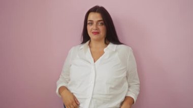 Genç bir kadın beyaz bir gömlek giyerken pembe bir arka plan üzerinde ellerini işaret edip işaret ediyor.