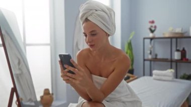 Kaplıcada dinlenen, kafasında bir havluyla akıllı telefon kullanan, huzur dolu bir spa günü boyunca huzur verici iç dekorasyonla çevrili bir sağlık merkezinde oturan bir kadın.