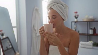 Kafasında havluyla spa odasında dinlenen bir kadın, huzur dolu bir dekoru ve sağlık merkezinde hafif ışıklandırmayla çevrili bir fincan çayın tadını çıkarıyor.