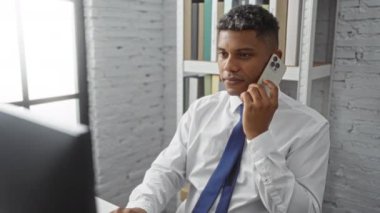 Ofiste telefonla konuşan genç bir adam. Yakınlarda bir bilgisayar ve arka planda kitap rafları var.