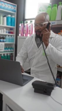 İçerideki raflarda çeşitli ürünlerle çevrili bir eczanede dizüstü bilgisayar ve telefonla çalışan eczacı.