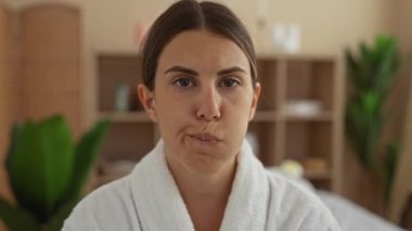 Kaplıca ortamında ağzını çalkalayan, esmer saçlı, beyaz bir bornoz giyen, rahat görünen, arka planda bitki ve raflarla kapalı bir sağlık odasında