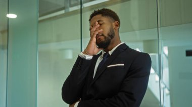 Takım elbiseli Afro-Amerikan bir adam eli yüzü üzerinde evde duruyor, modern ofis ortamında stres veya hüsranı tasvir ediyor..