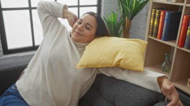 Rahat, orta yaşlı, İspanyol bir kadın rahat bir evde sarı bir yastıkla kanepede uzanıyor..