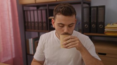 Sakallı genç bir İspanyol, dosyalar ve kutularla çevrili bir ofiste kahvesini yudumluyor..