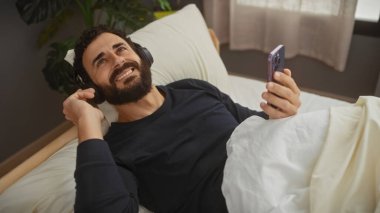 Sakallı gülümseyen bir adam, kulaklık takıyor, akıllı telefonu tutarken yatakta dinleniyor, yatak odası düzeninde..