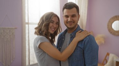 Çift gülümsüyor ve geniş bir pencereden içeri giren yumuşak doğal ışıkla ev ortamında kucaklaşıyorlar.