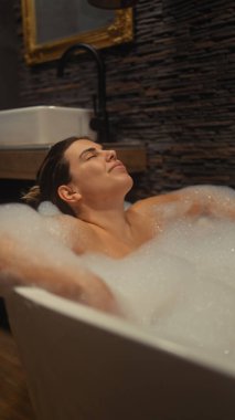 Huzurlu genç bir kadın modern bir banyo içinde lüks bir köpük banyosunun tadını çıkarıyor..