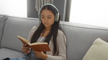 Evde rahat bir oturma odasında kulaklıkla müzik dinlerken kitap okuyan Çinli genç bir kadın.