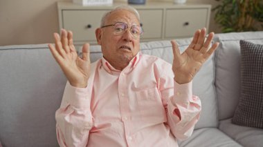 Gri saçlı ve pembe gözlüklü yaşlı bir adam şaşkın bir ifadeyle oturma odasındaki bir kanepede oturur ve ellerini kaldırır..