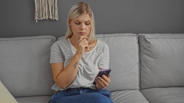 Oturma odasındaki gri koltukta oturan sarı saçlı genç kadın akıllı telefonuna dikkatle bakıyor.