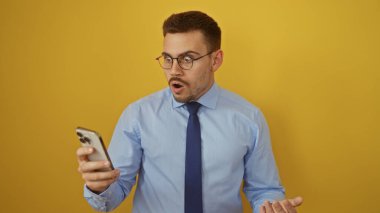Mavi tişörtlü ve gözlüklü İspanyol bir adam izole edilmiş sarı bir arkaplanda telefonuna bakarken şaşırdı..