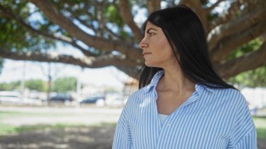 Güneşli bir günde ağaçlarla ve yeşilliklerle dolu bir parkta duran esmer saçlı, güzel İspanyol bir kadın..