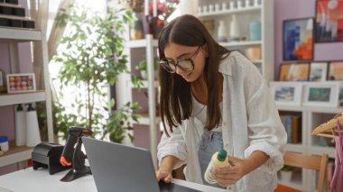 Dekorasyon malzemelerinin raflarıyla çevrili bir ev dekorasyonu dükkanında çalışan genç bir kadın elinde bir ürünü tutarken ve gözlük takarken dizüstü bilgisayarına odaklanmış..