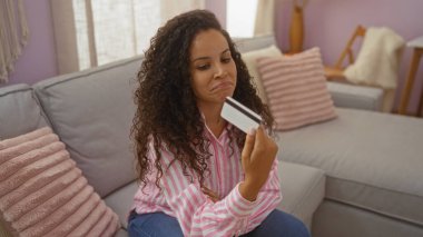 Kanepede oturan bir kadın, kredi kartıyla, rahat bir oturma odasında karışık duygular sergiliyor, modern bir iç dekorasyon ve sıradan bir atmosfer sergiliyor..