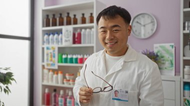 Yakışıklı genç Çinli bir adam gülümsüyor ve iyi organize edilmiş bir eczanede kadehler tutuyor, raflarda düzenli ürünler sergiliyor, profesyonel bir eczane ortamını yansıtıyor..