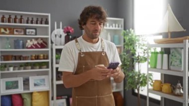 Genç adam çeşitli ürün ve bitkilerle dolu bir ev dekoratörü dükkanında akıllı telefon kullanıyor.