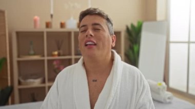 Beyaz bornozlu genç adam, sakin bir spa ortamında gözleri kapalı bir şekilde dinleniyor. İçerideki bitkiler ve hafif ışıklandırmayla, bir anlık sakinlik ve kendine bakıcılık izlenimi uyandırıyor.