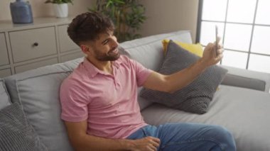 Sakallı genç İspanyol adam selfie çekiyor ve modern evin oturma odasında pembe gömlekli, koltukta rahatlamış bir şekilde akıllı telefon kullanıyor.
