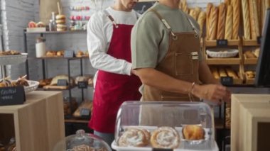 İki adam, ekmek ve hamur işleriyle çevrili bir dükkânda önlük giyip birlikte çalışıyorlar.