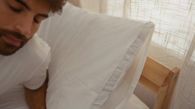 Sakallı genç adam beyaz yastıklar ve perdelerle güneşli bir yatak odasında huzur içinde uyuyor.