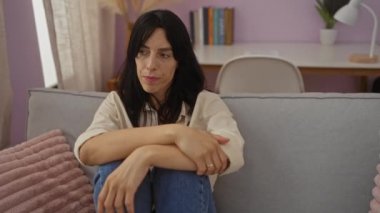 Rahat bir oturma odasında, koltukta oturan, açık mor duvarlı, beyaz gömlekli, mavi kotlu, yanında yumuşak bir yastıkla dalgın dalgın ileriye bakan bir kadın..