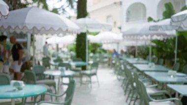 Lüks bir teras restoranında bulanık adamlar. Odakları çözülmüş bokeh arka planı ve yemyeşil şemsiyelerle çevrili boş masaları var.