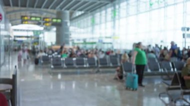 Odaklanamayan yolcularla dolu havaalanı terminali, modern mimari bir çatı yapısı altında, bavullarıyla bekleyen insanların yoğun bir sahnesini gösteriyor..