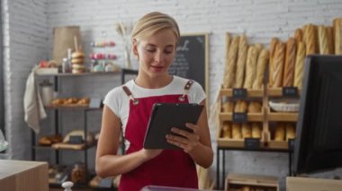 Fırında çalışan genç bir kadın, tablet kullanarak, çeşitli ekmek ve hamur işleriyle çevrili, kapalı bir dükkan ortamında, müşteriler için profesyonel ve çekici bir ortam sergiliyor.