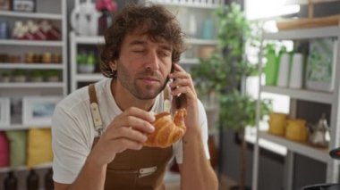 Genç bir adam cep telefonuyla konuşurken kruvasan yiyor. Ev dekorasyonunun şık olduğu bir mağazada.