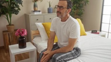 Yatak odasındaki bir yatakta oturan İspanyol bir adam, düşünceli bir yüz ifadesiyle, rahat bir evin içinde çiçekler ve bitkilerle çevrili..