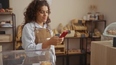 Çizgili gömlekli, kahverengi önlüklü, kahverengi saçlı genç bir kadın ekmek ve hamur işleriyle dolu bir fırında kırmızı bir telefon tutarken gülümsüyor.