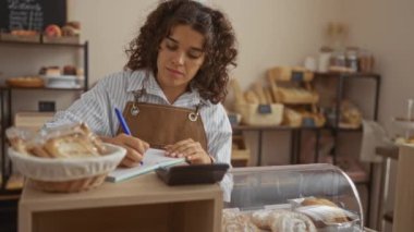 Ekmek ve hamur işleriyle çevrili fırın tezgahında deftere yazan genç bir kadın.