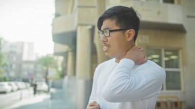 Gözlüklü genç Asyalı bir adam şehrin arka planında boyun ağrısı çekiyor..