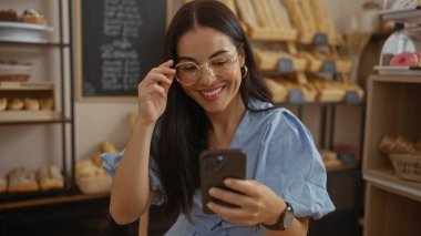 Fırındaki esmer kadın, gülümsüyor ve akıllı telefon kullanıyor, gözlüğünü ayarlarken, etrafı ekmek ve hamur işleriyle çevrili..