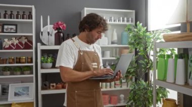 İç mekanı aydınlatan dekoratif bir mağazada dizüstü bilgisayar, mumlar, konteynırlar, raflar, bitkiler ve doğal ışık ile çevrili genç adam.