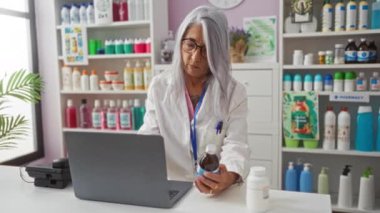 Eczanedeki ilaç şişesini inceleyen bir kadın dizüstü bilgisayarında çalışırken etrafını renkli ilaç ürünleriyle doldurmuş.