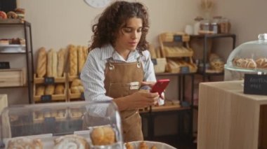 Genç bir kadın fırında akıllı telefon kullanıyor. Etrafı ekmek ve hamur işleriyle çevrili.