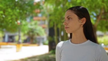 Güzel, genç, esmer bir kadın şehir parkının arka planında yeşil ağaçlar ve binalarla dışarıda duruyor.