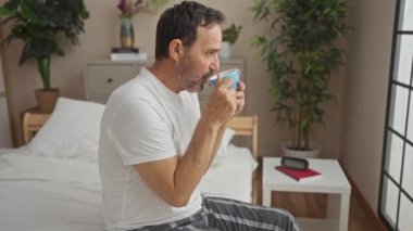 Orta yaşlı bir adam rahat bir yatak odasında kahvesini yudumluyor. Çiçekler ve modern dekorasyonla. Komodinin yanında oturmuş, huzurlu bir sabahın tadını çıkarıyor..