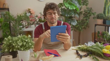 Sakallı İspanyol adam, kapalı çiçekçide tablet kullanıyor. Etrafı bitki ve buketlerle çevrili..