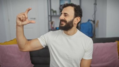 Sakallı, orta yaşlı bir adam, modern bir apartman dairesinde gülümseyen bir ifadeyle küçük bir mesafe jesti yapıyor..