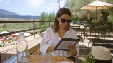 Genç bir kadın güneşli Mallorca, İspanya 'da bir masada kitap okuyor, manzara manzaralı, güneş gözlüğü takıyor ve günlük beyaz bir gömlek giyiyor..