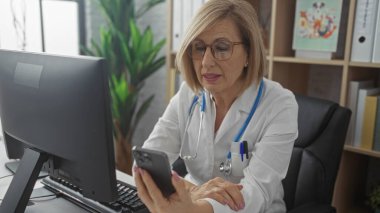 Olgun, kadın, doktor bir klinikte akıllı telefon kullanıyor, bilgisayarlı bir masada oturuyor, gözlük takıyor ve stetoskop takıyor, parlak, düzenli bir iç çalışma alanında..
